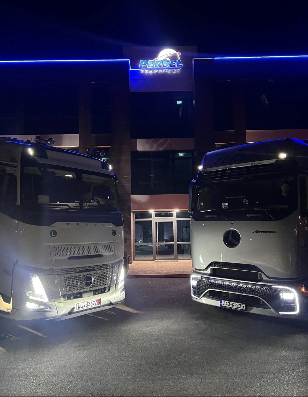 Dva kamiona Persel Transport sa upaljenim farovima noću ispred osvijetljene zgrade firme
